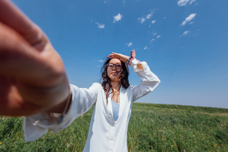 A cute young Asian woman with glasses smiles against a background of blue sky and green dense grass. A charming Korean girl in casual clothes is relaxing and laughing in nature in the outdoors.の写真素材