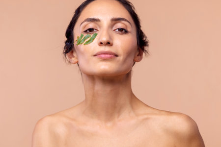 Studio portrait of a young woman with leaves under her eye on a beige isolated background. Natural eco-friendly cosmetics. Taking care of clean and delicate facial skin. Cosmetology. Dermatology. Spa.の写真素材
