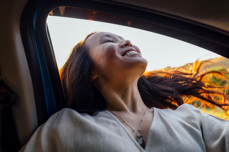 Young attractive Asian woman enjoys a ride in a automobile. Charming Korean sticks her head out of the car window and catches the wind. Cute girl having fun at the wheel. Beautiful sunset and nature.の写真素材