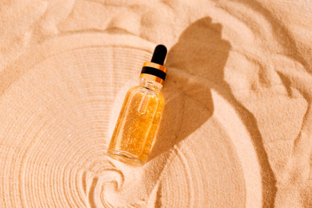 A mock-up glass bottle with a pipette with an intensive moisturizing concentrate with hyaluronic acid and vitamins on a background of sand. Gold glitter serum for the face with peptides and collagen.の写真素材