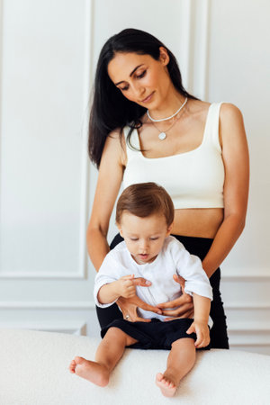 A beautiful young woman in a white top and black leggings holds her little son in a white shirt and black shorts. An adorable mom hugs her sweet baby. Family look.の写真素材