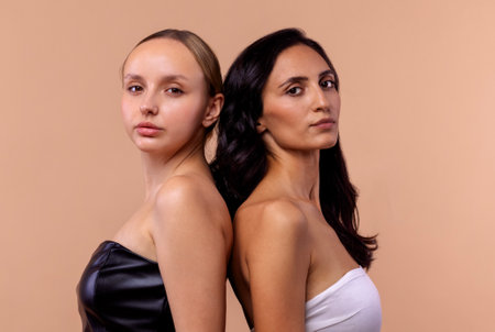 Two young diverse women wearing tops looking confident at camera while posing together on beige background. Charming models with natural makeup and perfect skin. Diversity, beauty, friendship concept.の写真素材