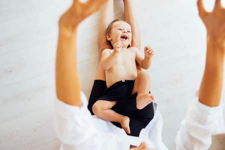 Close-up of a laughing little boy lying on his mom's legs. A sweet smiling toddler is having fun with his parent at home. A mother tickles her cute baby while sitting on the floor.の写真素材