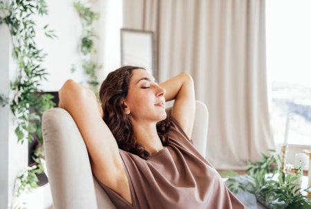 Young woman is sitting and relaxing with her arms behind her back and her eyes closed on the sofa at home. Cute curly-haired girl is resting on the couch in the living room. Modern cozy interior.の写真素材