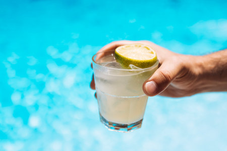 Young male hand holds a glass of a chilled drink with ice cubes. White cocktail with a lemon slice. Sunny day and reflection of rays in the blue pool water on background. Relaxation, vacation concept.の写真素材