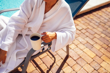 Young pretty woman in a white bathrobe lies on a sun lounger and holds a cup of coffee. Relaxed caucasian female in sunglasses and a towel on her head sits at the poolside. Vacation and spa concept.の写真素材