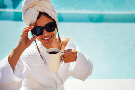 Young pretty woman in a white bathrobe by the swimming pool and holds a cup of coffee. Relaxed caucasian female in sunglasses and a towel on her head sits at the poolside. Vacation and spa concept.の写真素材
