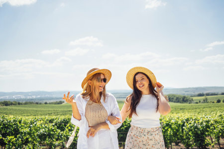 Two happy young women embrace and walk through the vineyard. White female friends in casual clothes and straw hats look at the landscape. Friendship and travel concept. Copy space.の写真素材