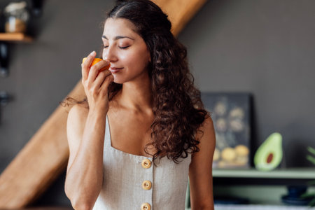 A young attractive woman closes her eyes and sniffs half an orange. A curly-haired smiling girl is eating an orange. A charming brunette enjoys the smell of citrus. Cozy and modern interior.の写真素材