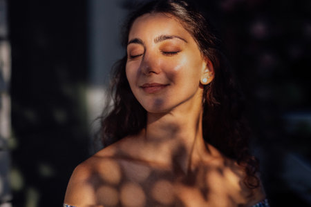 Close-up of a female portrait of a charming curly-haired young woman. An attractive brunette is smiling and her eyes closed outdoors in the park. Natural beauty. The sun and shade from the trees.の写真素材