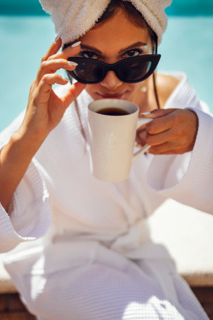 Young pretty woman in a white bathrobe by the swimming pool and holds a cup of coffee. Relaxed caucasian female in sunglasses and a towel on her head sits at the poolside. Vacation and spa concept.の写真素材