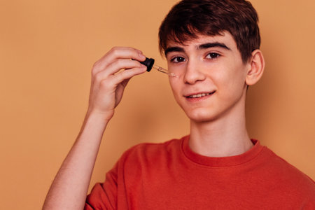 Teenage boy applying cleansing serum against acne and pimples on his cheek. Male teen in casual...