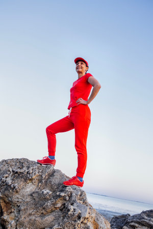 An attractive young woman in a red tracksuit stands on top of a mountain. A charming girl in a winning pose smiles and enjoys nature by the sea. A successful handsome fitness model.の写真素材