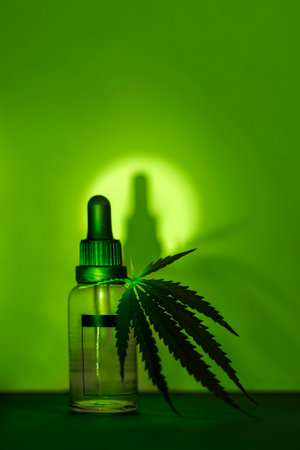 A mock-up glass transparent bottle with a pipette with a moisturizing concentrate with hyaluronic acid and leaf of cannabis. Facial serum with peptides and collagen with hemp. Green background.の写真素材