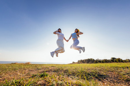 Two multiethnic friends in casual clothes and panama hats are jumping and laughing outdoors. Young cheerful women of different races have fun together in nature. Seascape. Freedom and friendship.の写真素材