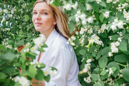Close up of a portrait of a young beautiful blonde woman in a white shirt with jasmine flowers. A charming girl smiles and poses at the camera in a garden or park. Romantic atmosphere and dreamy mood.の写真素材