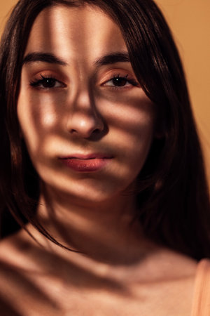 A close-up portrait of a thoughtful female teenager in the warm lighting of the room. A sweet beautiful girl with a shadow of leaves on her face. Beige background.の写真素材