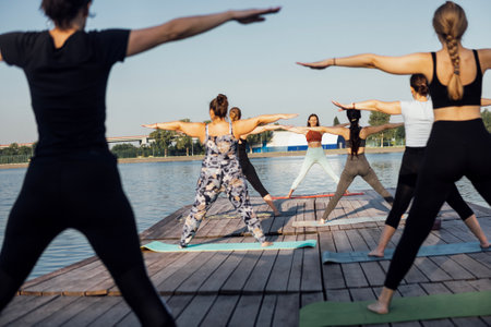 Morning yoga class outdoors with instructor near river in city. Young women stand with their arms...