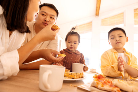 A happy Asian family is eating pizza in the kitchen at home. A young Korean couple and their cute children are having lunch. Dad, mom, daughter and son are eating a pie.の写真素材