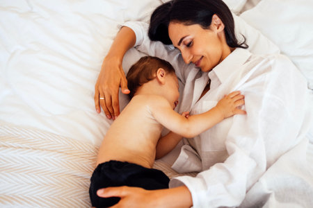 A cute baby sleeps with his attractive mother in a bed. A little boy falls asleep in the arms of his mother. A young smiling woman puts her sweet son to bed.の写真素材