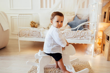 A cute little boy is riding a white wooden horse in his room. A sweet baby is playing in the nursery. A funny toddler in casual clothes is swinging on a gurney at home.の写真素材