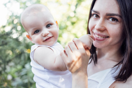 Close up of female portrait of young charming mother holds her cute baby in arms in garden. Pretty smiling infant with his attractive brunette mom in white clothes outdoors. Green trees on background.の写真素材