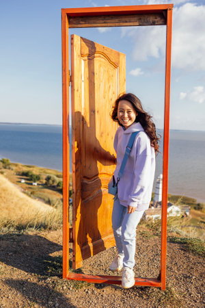 A young attractive Asian woman laughs and opens the door outdoors. A door on a hill and a beautiful seascape. A charming smiling Korean woman in casual clothes walks into the doorway.の写真素材