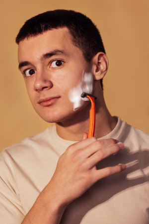 A close-up portrait of a teenager with foam on his cheek and holding a razor. A handsome guy in a T-shirt is shaving. Hygiene and skin care in adolescence. Isolated beige background.の写真素材