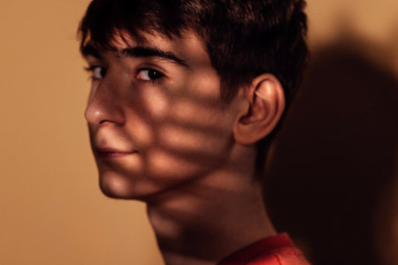 Close-up portrait of a male thoughtful teenager in warm indoor light. A pensive young man in profile with shadows of a leaf playing on his face. Beige background.の写真素材