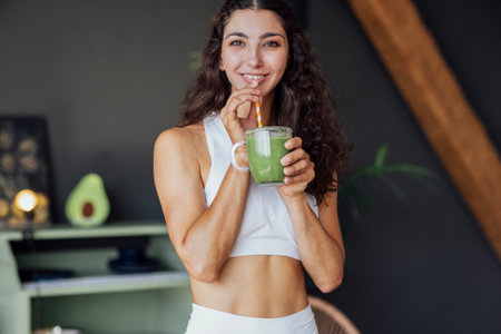 Attractive curly-haired girl in white top smiles and drinks green smoothie from transparent glass. Fit charming young woman in fitness suit drinks healthy drink at home. Modern and cozy interior.の写真素材