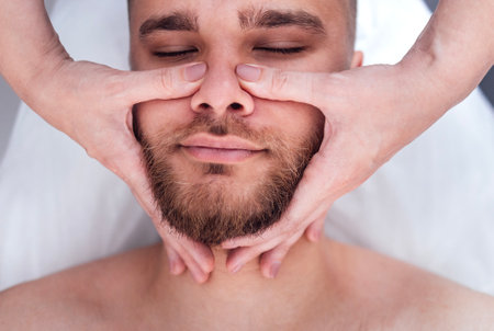 Close up portrait of a young attractive man getting facial massage. Masseuse hands on the male client chin. Brunette girl closed her eyes and enjoys a spa procedure in beauty salon.の写真素材