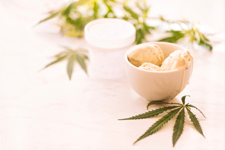 A hemp leaf is lying on a mock-up jars of ice cream on a white marble table. Green cannabis leaves on the background. Organic food and health.の写真素材