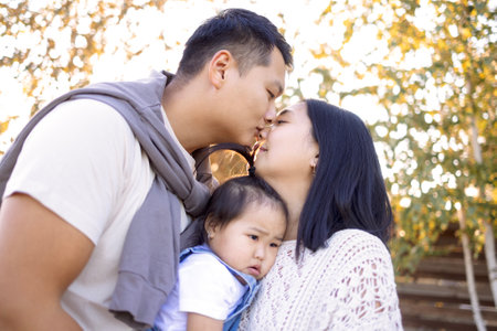 An Asian couple in love is kissing outdoors and holding their kid. Cute affectionate Korean parents in casual clothes hug their little daughter. A happy family. Mom and dad with their sweet baby.の写真素材
