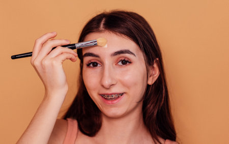 A female teenager puts a moisturizing mask or cream on her face. A pretty girl does makeup with a brush. Hygiene and skin care in adolescence. Isolated beige background.の写真素材