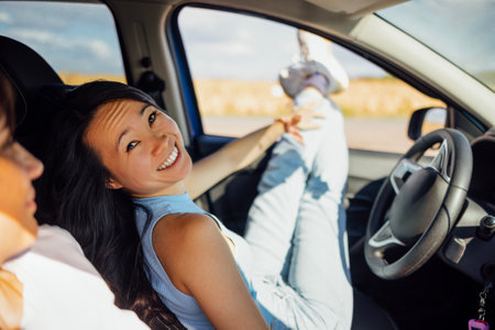 Young women look out of the car window and laugh. Smiling girls of different races travel and have fun on the road. Charming multiethnic friends enjoying nature. Journey, vacation and freedom concept.の写真素材