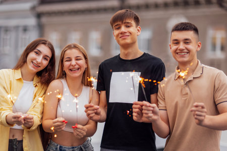 Two cute guys and two charming girls are holding sparklers and smiling. Friends in casual clothes have fun and laugh. Four teenagers celebrate a date or holiday. Happy teens on a rooftop party.の写真素材