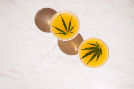 Two elegant long-stemmed glasses with green cocktails with cannabis leaves on the light marble...