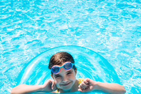 A little girl with an inflatable circle jumped into the pool. A cute kid in a swimsuit and swimming glasses is swimming in clear water. The view from the back. Vacation, weekend and relax concept.の写真素材