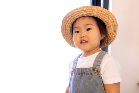A close-up portrait of a cute little Asian girl. Funny Korean toddler in a straw hat, white T-shirt and denim jumpsuit at home. Copy space.の写真素材