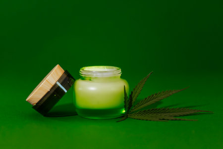 A mock-up of a transparent glass bottle with a cream with moisturizing concentrate, hyaluronic acid with lid and hemp leaf. Facial serum in a jar with peptides and collagen with hemp. Green backgroundの写真素材