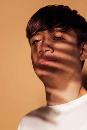 Close-up portrait of a male thoughtful teenager in warm indoor light. A pensive young man in profile with shadows of a leaf playing on his face. Beige background.の写真素材
