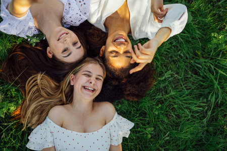 Three cute female teens of different ethnicities in white elegant dresses lie on grass. Smiling girls have great time together on meadow. Funny caucasian and mixed race teenagers laugh and enjoy life.の写真素材