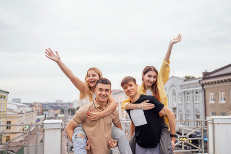 Cute girls in casual clothes ride on the backs of handsome guys on the roof of the house. Two young...
