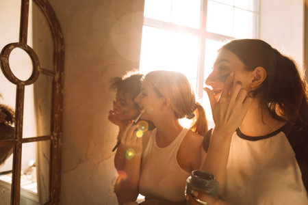 Three charming teenagers of different races apply cosmetic masks to their faces in front of a mirror. Multiethnic female friends take care of their skin. Cute funny students have fun together at home.の写真素材