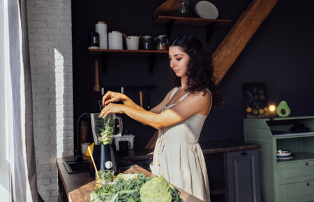 A beautiful curly-haired brunette in a linen sundress is cutting greens in the kitchen. A young attractive smiling vegetarian woman makes a green smoothie at home. Modern cozy sunny interior.の写真素材