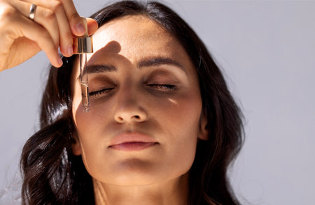 Close-up of charming woman with her eyes closed holding pipette with regenerating serum with hyaluronic acid. Attractive girl with beautiful healthy skin and moisturizing anti-aging facial essence.の写真素材