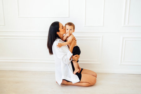 A young mother is sitting on the floor in the living room and holding her cute baby in her arms. An...