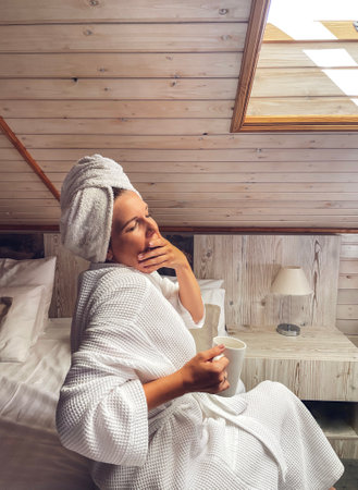 Young woman in a white bathrobe and a towel on her head is holding a cup. Relaxed caucasian female is drinking tea or coffee near an open skylight in a modern interior. Vacation and spa concept.の写真素材