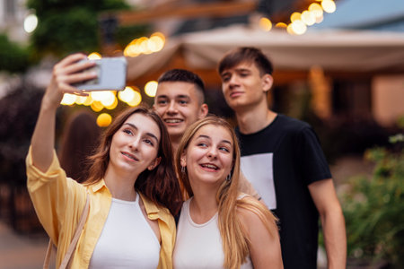 Four cute teenagers taking a selfie. Funny friends takes a picture of themselves on the phone outdoors. Smiling boys and girls sitting on the street. Happy teens enjoying vacation together.の写真素材