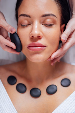 Close-up of a portrait of a young woman with her eyes closed receiving a facial massage with hot stones. A beautiful girl lies and relaxes during a spa procedure. Taking care of yourself concert.の写真素材
